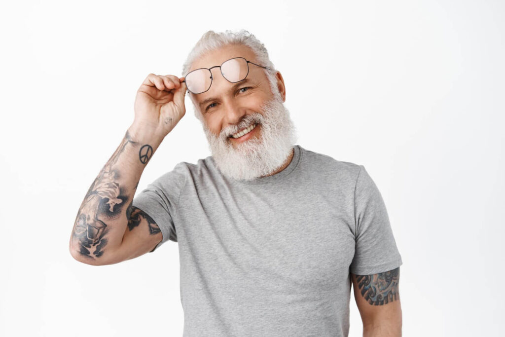 Homem sênior de barba branca e tatuagens sorrindo ao ajustar óculos, representando a longevidade da prótese protocolo na Clínica Dahdal Aoun.