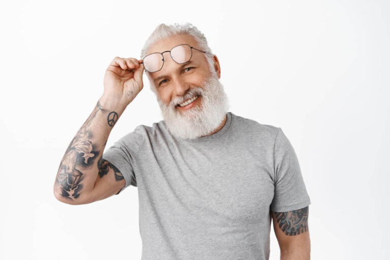 Homem sênior de barba branca e tatuagens sorrindo ao ajustar óculos, representando a longevidade da prótese protocolo na Clínica Dahdal Aoun.