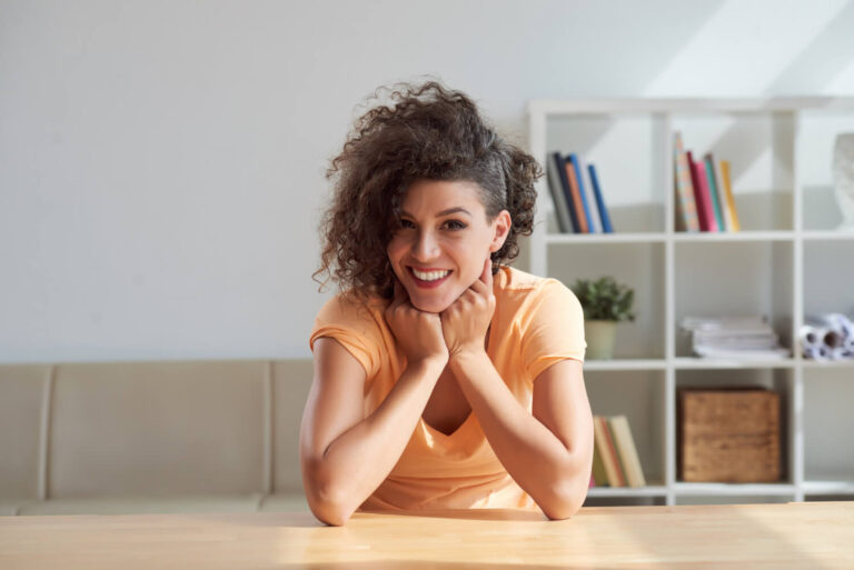 Mulher sorridente com cabelos cacheados apoiada em uma mesa de madeira clara. Transforme seu sorriso com facetas em resina em Moema.